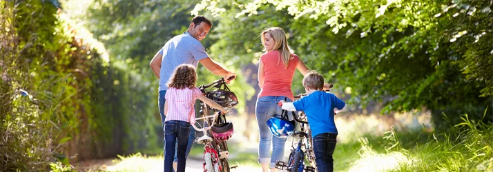 A family of four walks with bicycles along a sunlit path surrounded by greenery.